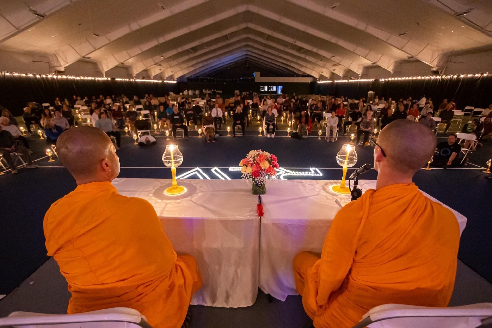 Two monks seated at a draped table on an indoor court, with a full house of guests behind them.