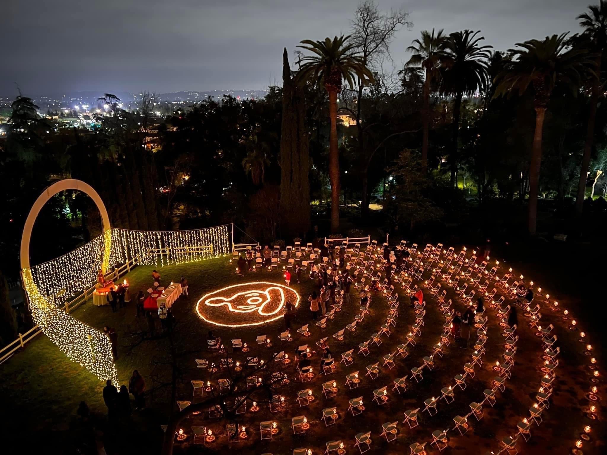 Seen from above at night: rings of candles and chairs across a hillside lawn beside a lit archway.