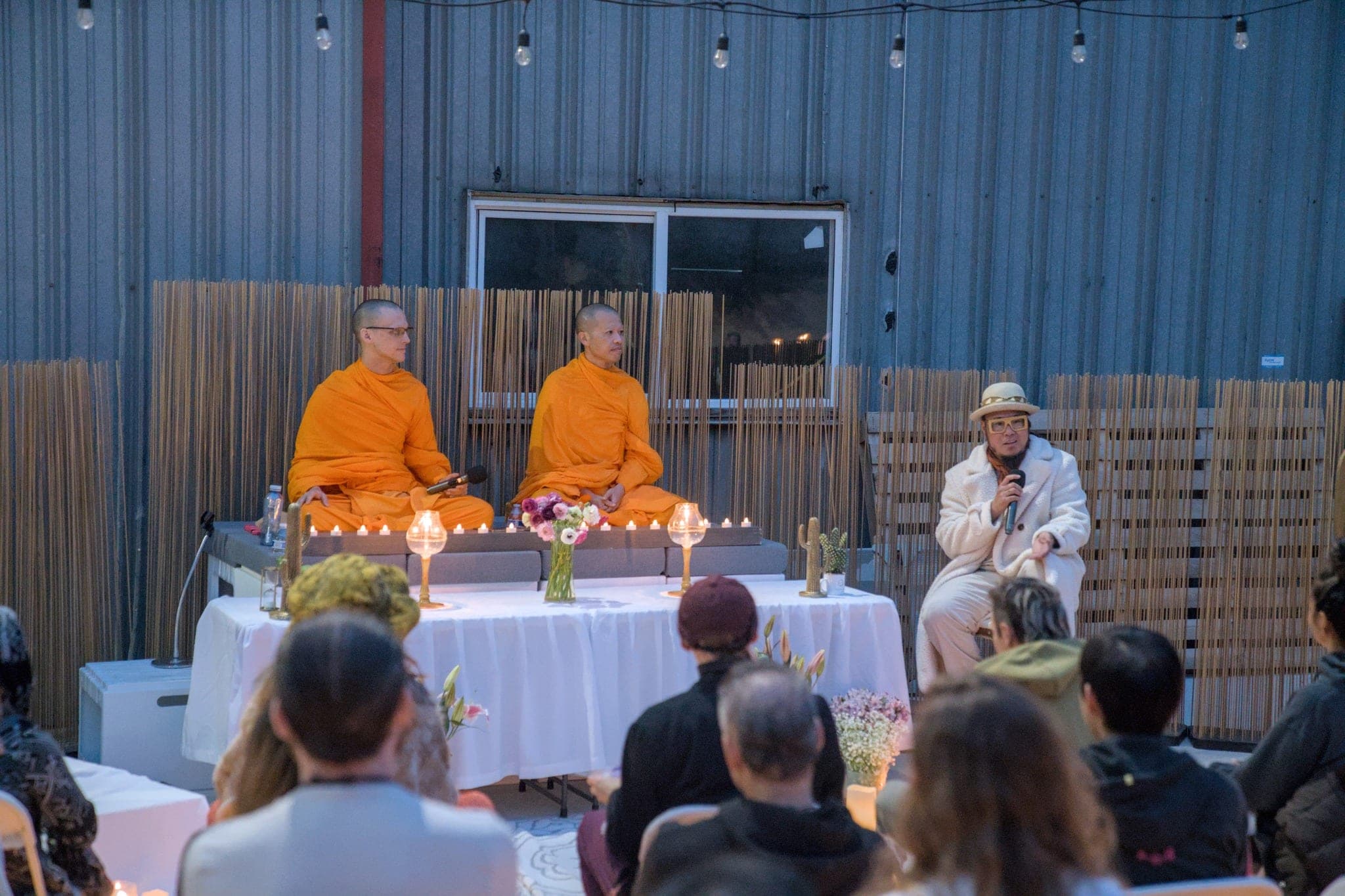 Three monks at a draped table beside a speaker, seen over the heads of the seated audience.