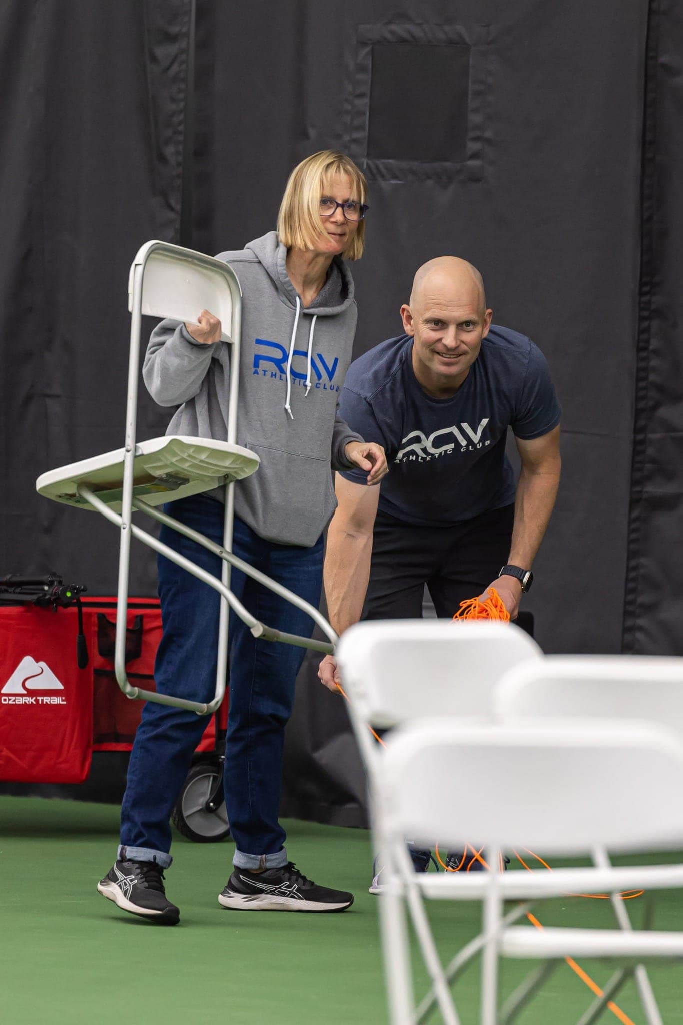 Two volunteers carrying folding chairs across an indoor court while setting up.