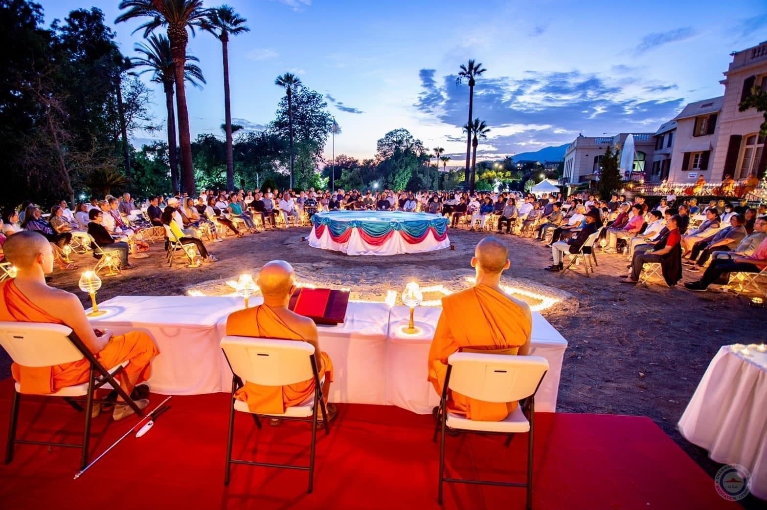 Monks seated at a draped table beside a pool at dusk, with guests seated all the way around it.