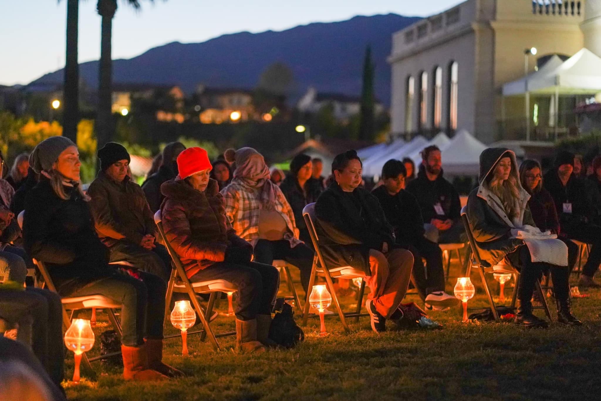 Guests seated on a lawn at dusk in coats and blankets, a candle lamp at each chair.